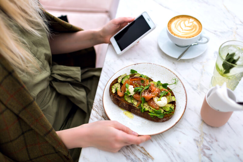 woman eating her tasty brunch of a toast, avocado and eggs with coffee, table top marble view, enjoying breakfast, looking at the mobile, hand holding mobile