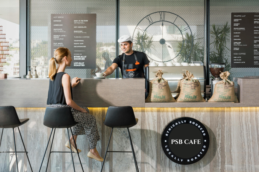 Barista serving coffee at PSB Café with menu and coffee bags in the background.